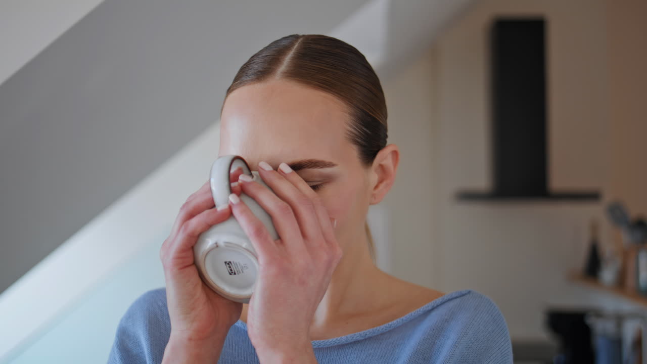 Portrait woman drinking tea cup looking at camera in morning sunlight interior