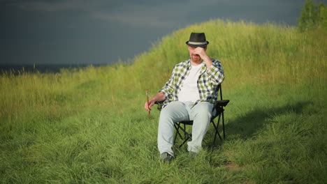 a man wearing a checkered shirt and jeans sits comfortably in a chair in a sunny grassy field, adjusting his hat with one hand while holding a paintbrush in the other