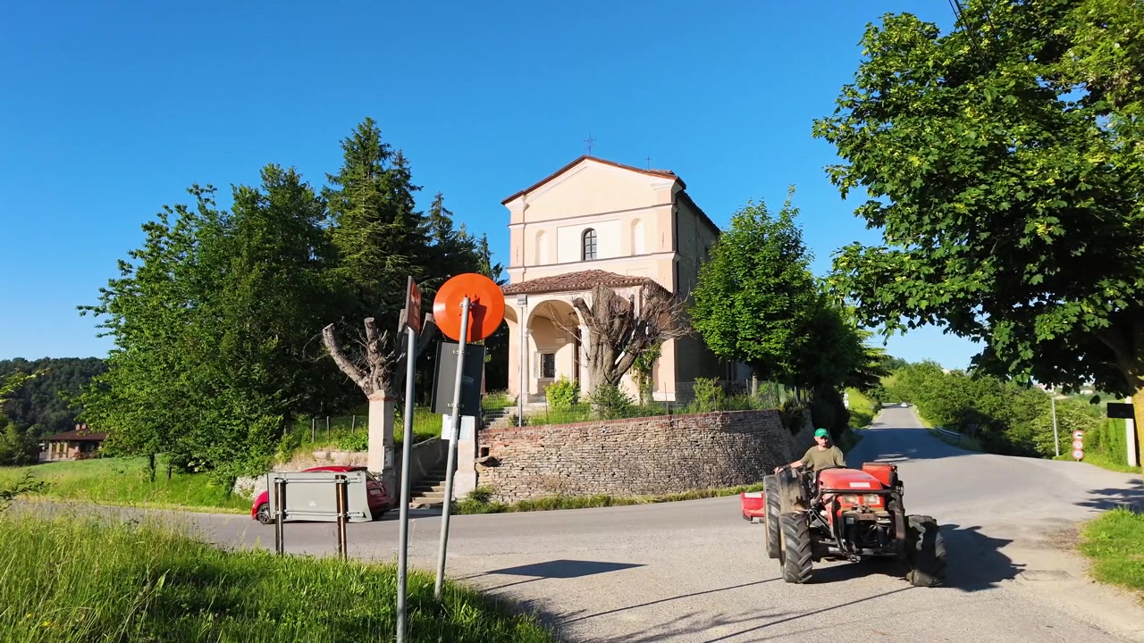 Un trattore rosso percorre una strada di campagna assolata passando accanto a una chiesa tradizionale italiana.