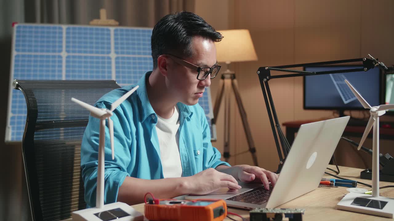 작은 풍력 터빈을 가진 아시아인 남자가 사무실에서 태양전지 에 노트북으로 타이핑하고 있습니다.