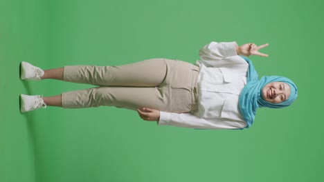 녹색 스크린 배경 스튜디오에 서있는 동안 카메라에 평화의 몸짓을 보여주고 미소 짓는 아시아 무슬림 여성의 몸이 가득 차 있습니다.