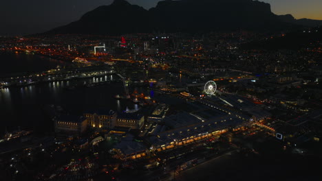 Blick-Aus-Dem-Hohen-Winkel-Auf-Die-Uferpromenade-Mit-Yachthafen-Bei-Nacht.-Kippen-Sie-Die-Stadt-Und-Die-Silhouette-Des-Bergrückens-Im-Hintergrund-Nach-Oben.-Kapstadt,-Süd-Afrika