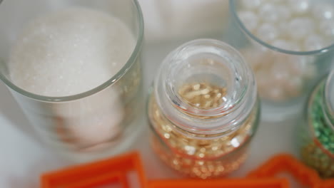 primer plano de cristales de azúcar espumosos en taza de vidrio junto a cuentas decorativas de oro almacenadas en pequeños frascos de vidrio, arregladas cuidadosamente para hornear y decorar postres, con cortadores de galletas naranjas visibles