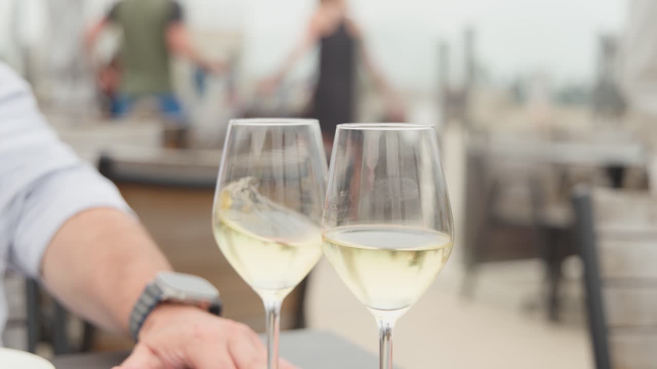 Two hands clink white wine glasses outdoors, soft daylight, shallow focus, relaxed coastal atmosphere