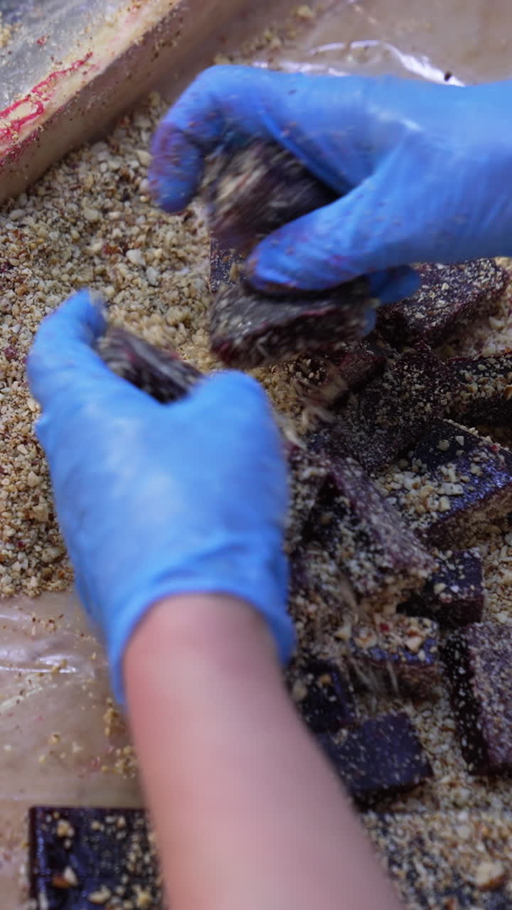 Dark marmalade pieces being coated with crushed nuts. Worker's hands mix up sweet desserts with ground walnuts. Close up. Vertical video