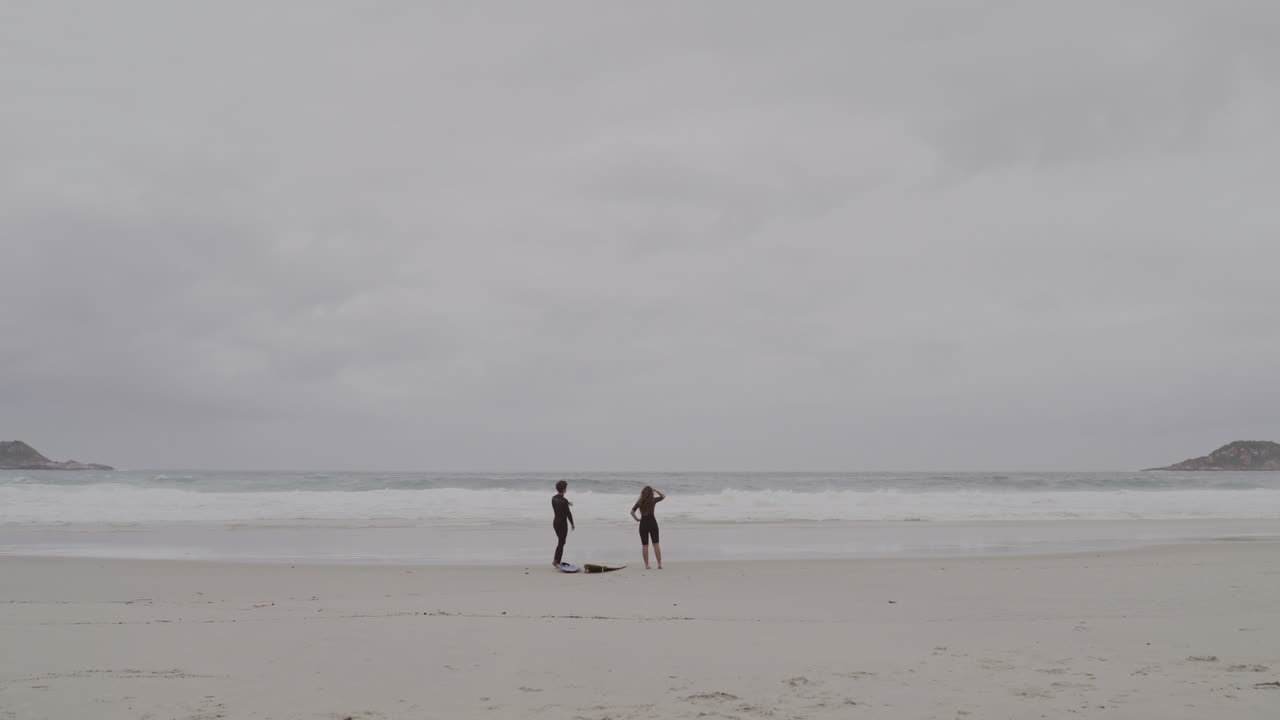 Two Surfers Standing on a Vast Beach Under an Overcast Sky