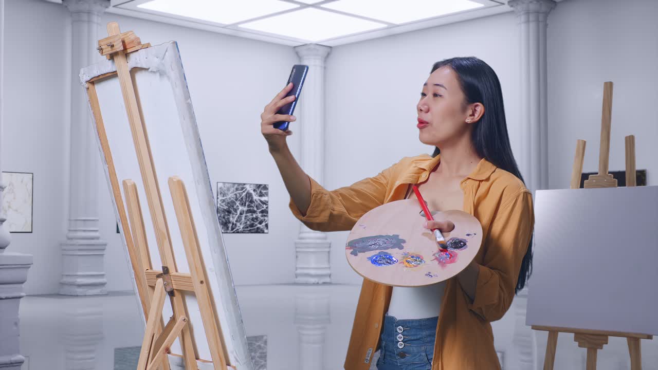 아시아 여성 예술가 가 갤러리 에서 오일 페인트 와 브러쉬 로 캔버스 에 그림 을 그리는 동안 스마트폰 으로 비디오 통화를 하고 있다