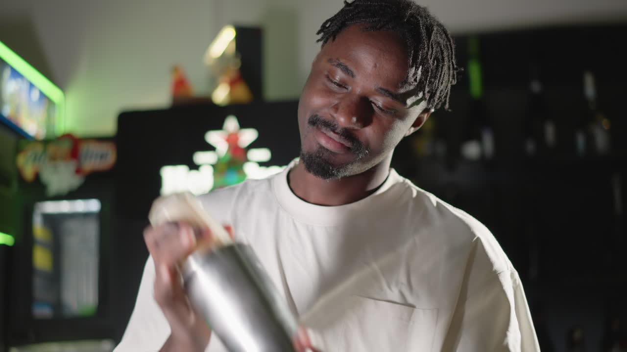 Bartender smiling while shaking cocktail shaker, preparing drink with focus. Bright bar ambiance with blurred bottles in background, perfect for videos showcasing cocktail making, mixology skills, and bartenders at work