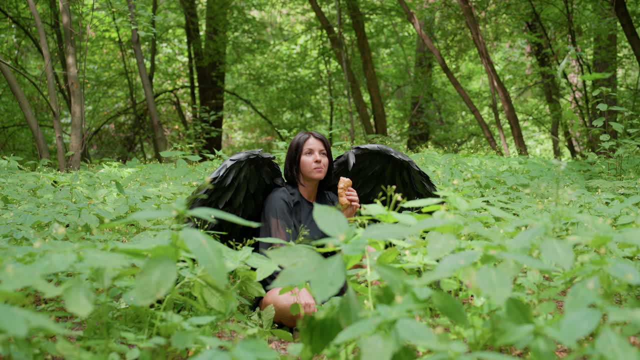 Mysterious woman dressed as dark goddess with black wings squats among green plants, eating pastry and sipping drink from flask cover, surrounded by lush forest atmosphere filled with calm