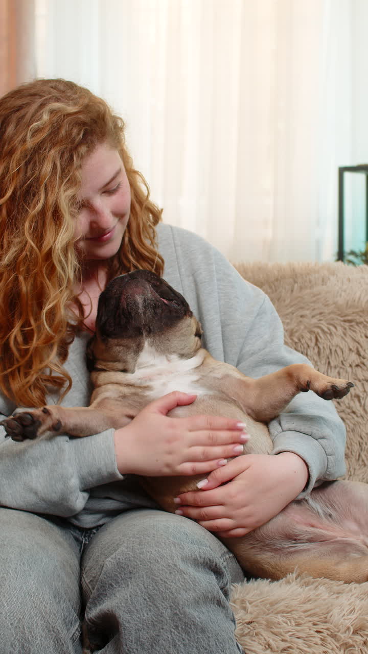 Young woman gently petting pug dog on home sofa with affection and calm expression in quiet moment
