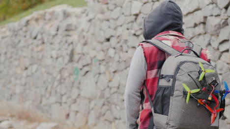 a man with a backpack behind him and a sports drone walks along a stone wall