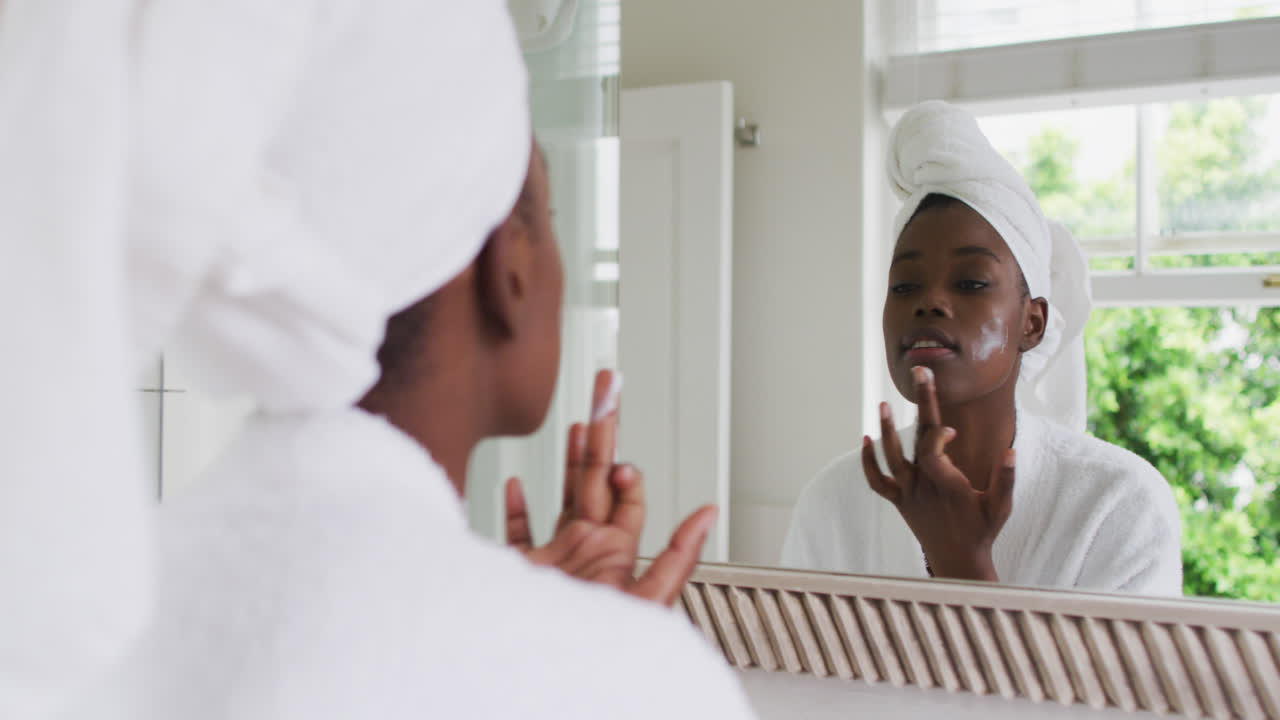African american woman in bathrobe applying face cream while looking in the mirror at home