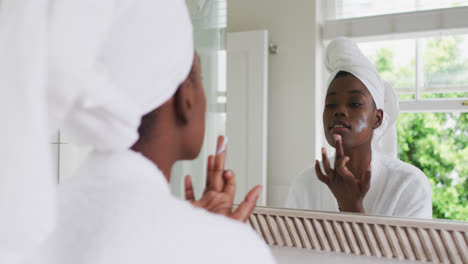 african american woman in bathrobe applying face cream while looking in the mirror at home