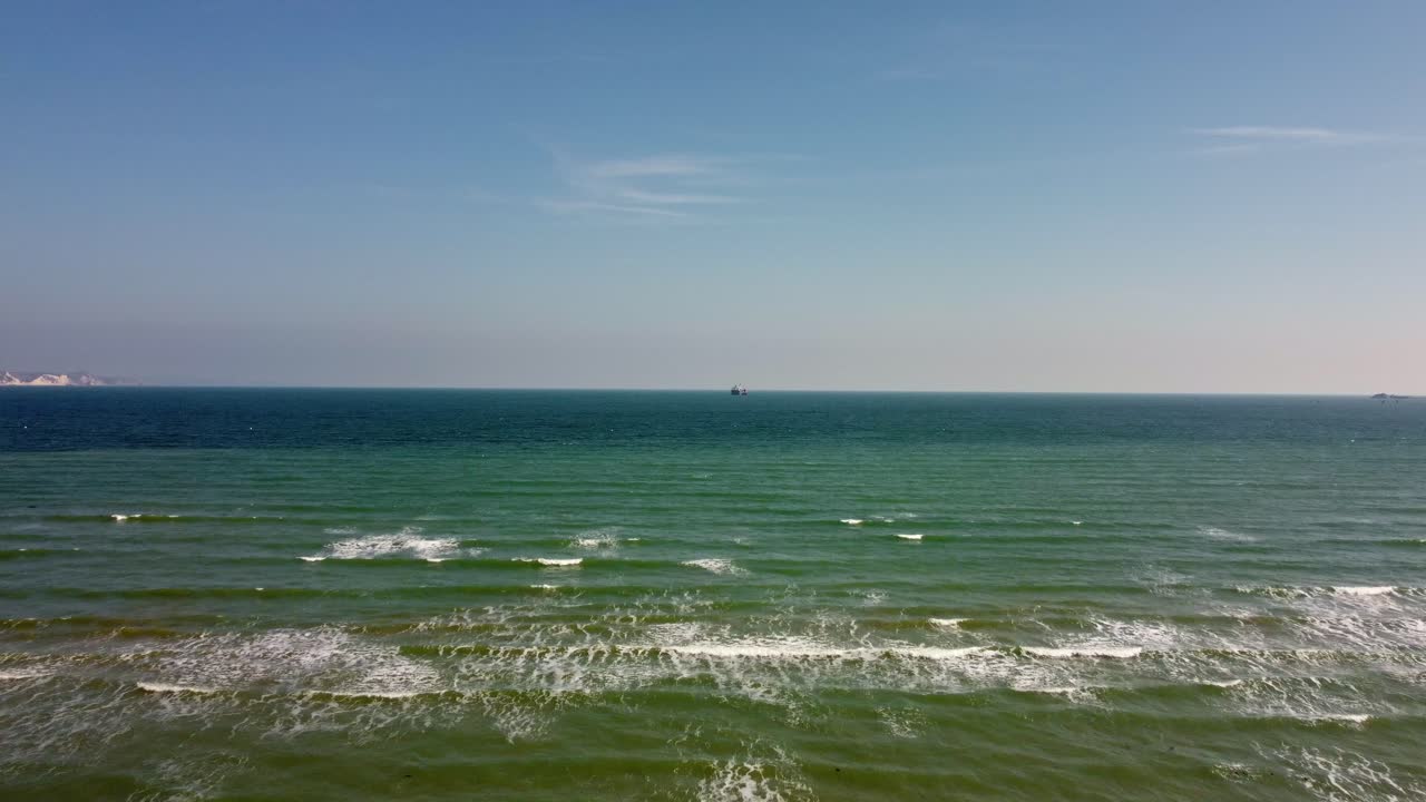 Drone video from Weymouth beach moving up and out to the sea on a sunny day with a commercial ship near the horizon.