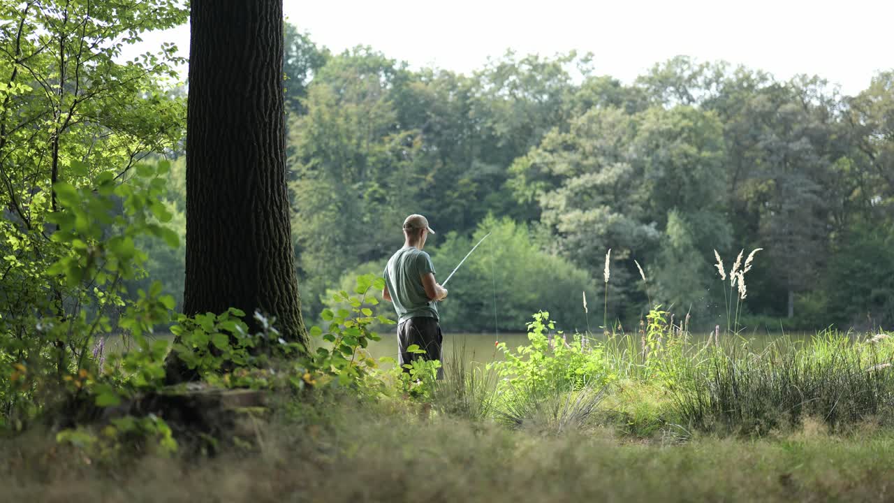 남성  ⁇ 시 ⁇  은 풀 과 나무 사이 에 흐린 호수 해안 에 서서  ⁇ 시대 로 물고기 를 잡고 있다