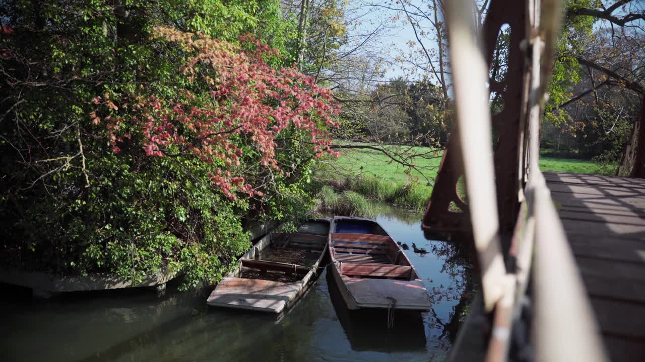 Two old wooden boats rest by the riverside under colorful spring foliage, partially shaded by a bridge