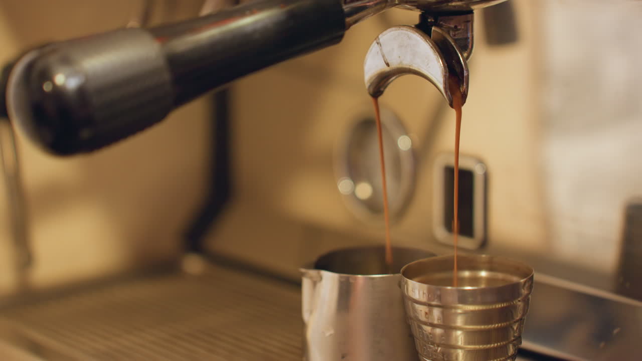 Primo piano di un caffè espresso che si versa in un bricco di metallo, crema ambrata calda che scorre dal portafiltro in un misurino, vassoio in acciaio inossidabile testurizzato, illuminazione da caffè vintage, atmosfera lenta e accogliente.