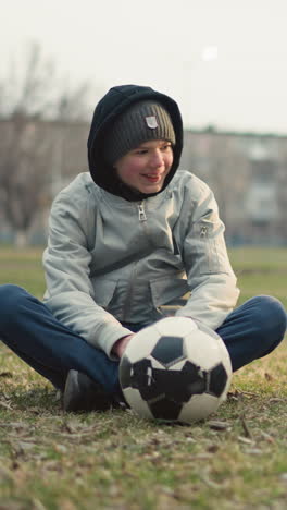 two boys seated on a grassy field, having a discussion and smiling, the boy in a black outfit focuses his attention on the one in a gray top who has a soccer ball in front of him