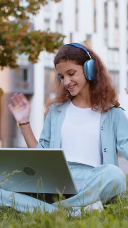 child girl making video laptop webcam conference call with friends or family sits on grass in park