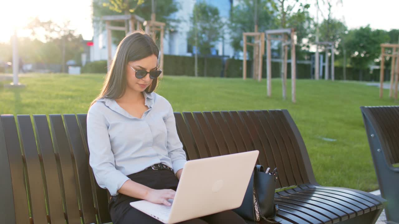 una mujer de negocios está sentada en un banco al aire libre y usando una computadora portátil para trabajar en la oficina