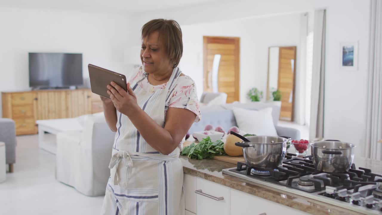 feliz mujer mayor afroamericana preparando comida en la cocina, usando una tableta y sonriendo