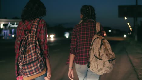 two young women hitchhikers stand on the sidelines holding their backpacks and ask to stop the passing cars in the night in summertime