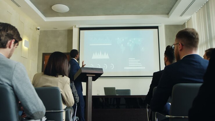 Camera zoom out on african american businessman talking on a podium in a conference room and showing some charts and graphics on the big screen