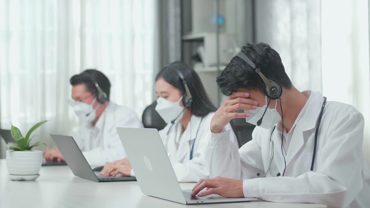 A Man Of Three Asian Doctors With Stethoscopes In Headsets And Masks Working As Call Center Agent Is Tired Due To Working While His Colleagues Are Speaking And Typing During A Call At The Office