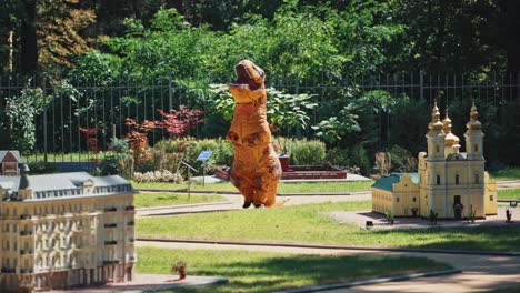 funny dino in the toy city outdoors. person in a big orange blow-up suit of a dragon walking in the park among small buildings.