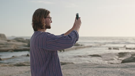 retrato de un hombre geek con gafas tomando fotos en la playa usando un teléfono