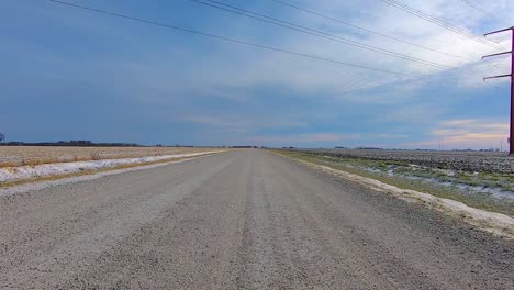 Rear-POV-while-driving-down-a-rural-gravel-road-past-harvested-fields-and-light-snow-covered-ditches