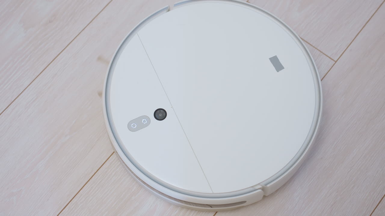 Close up of white robotic vacuum cleaner moving away on wooden floor near person seated wearing black socks, highlighting smart cleaning technology, domestic lifestyle automation