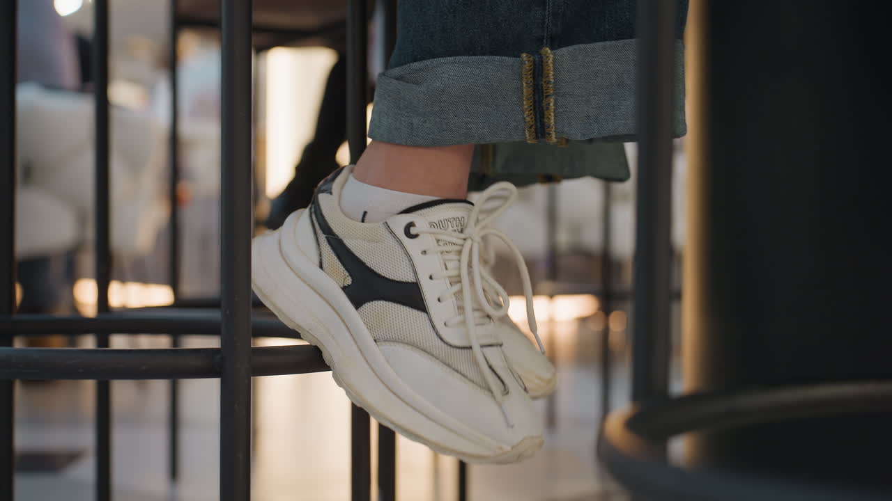 Primer plano de zapatillas deportivas sobre un taburete metálico, suela blanca gruesa y banda de rodadura texturizada, dobladillo de mezclilla enrollado que muestra un toque de estilo, sutil rebote y movimiento, ambiente de tienda con luz cálida ambiental, enfoque en el material