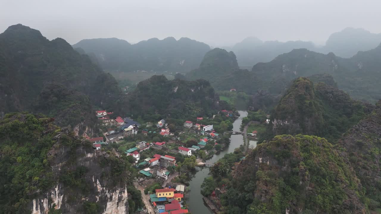 Aerial perspective over Trang An, Ninh Binh, Vietnam, revealing limestone karst mountains, a winding river, and a village with traditional houses surrounded by lush greenery and misty hills
