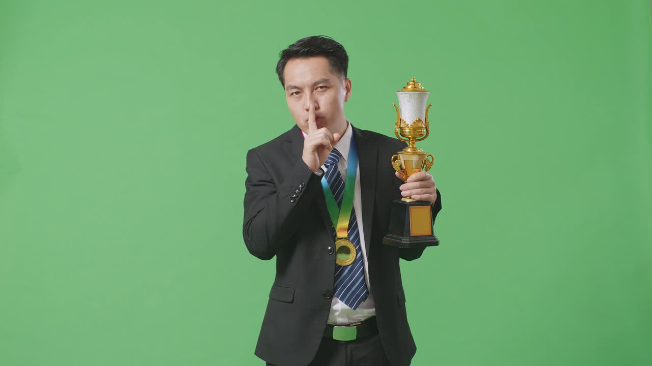 슈트와 타이를 입은 아시아 사업가, 금메달과 트로피를 들고, 조용한 제스처를 하고, 스튜디오의 녹색 스크린 배경에서 첫 번째 우승자로서 승리하는 것이 비밀이라고 말합니다.