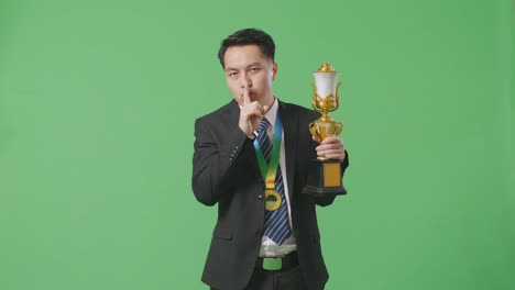 hombre de negocios asiático en traje y corbata con una medalla de oro y trofeo haciendo un gesto de silencio diciendo que es un secreto para ganar como el primer ganador en el fondo de la pantalla verde en el estudio