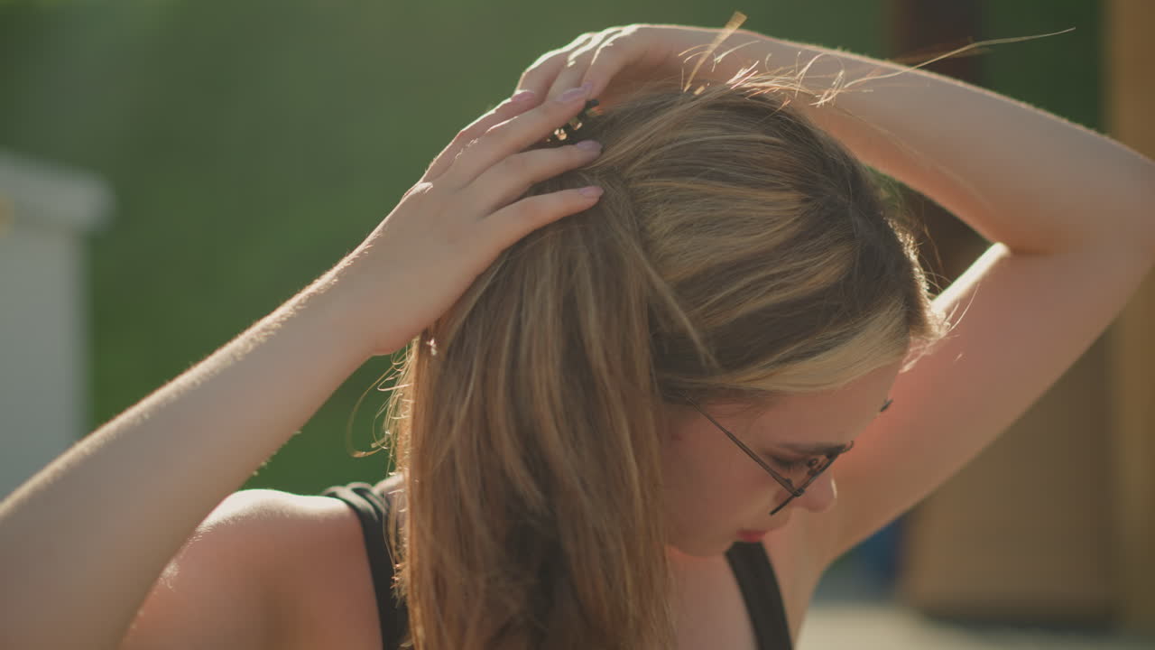 dama ajusta su cabello como el viento sopla a través de él mientras usa gafas de sol al aire libre, el sol irradia en ella, y un fondo de vegetación borrosa