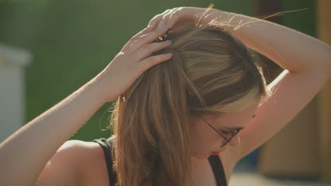 lady adjusts her hair as wind blows through it while wearing sunglasses outdoors, the sun radiates on her, and a blurred greenery background