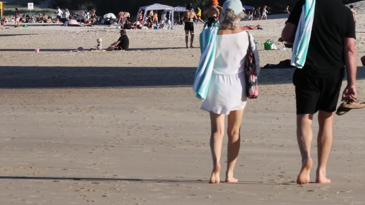 A couple walks along a sandy beach, surrounded by beachgoers enjoying a sunny day.