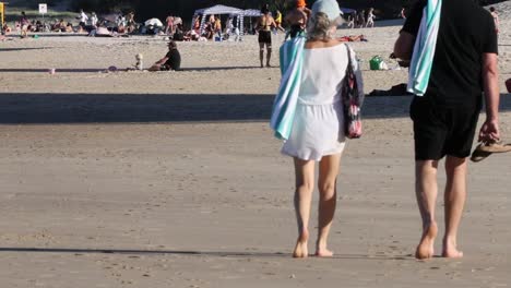 a couple walks along a sandy beach, surrounded by beachgoers enjoying a sunny day.