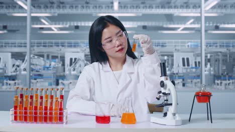 mujer científica asiática mirando el líquido naranja en el tubo de ensayo cuidadosamente mientras trabaja con el microscopio en el laboratorio, trabajando en el laboratorio analizando muestras tubo de ensaio