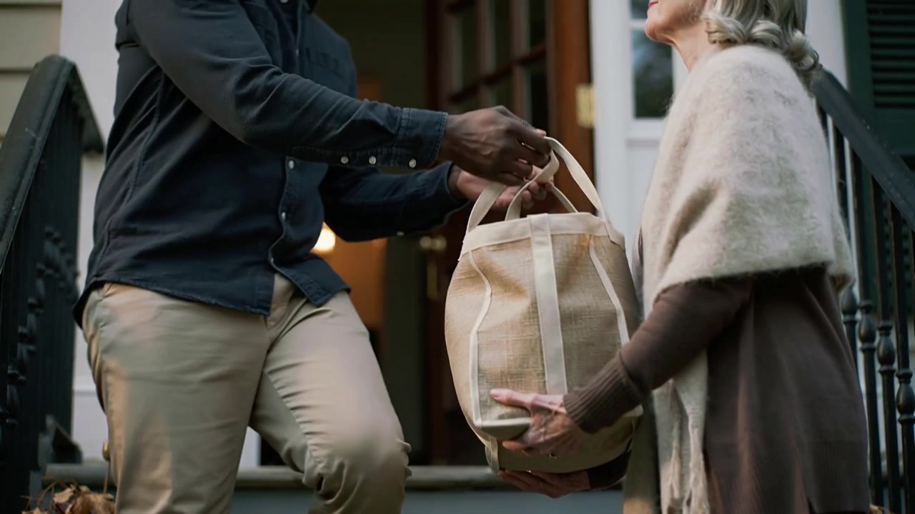 Handing tote, courier stepping from open door onto porch, delivering to senior woman wearing shawl