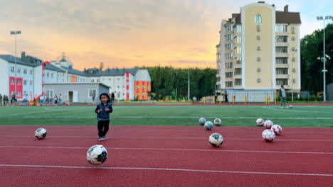 lovely toddler boy playing with football balls at the stadium. group of children training at backdrop.