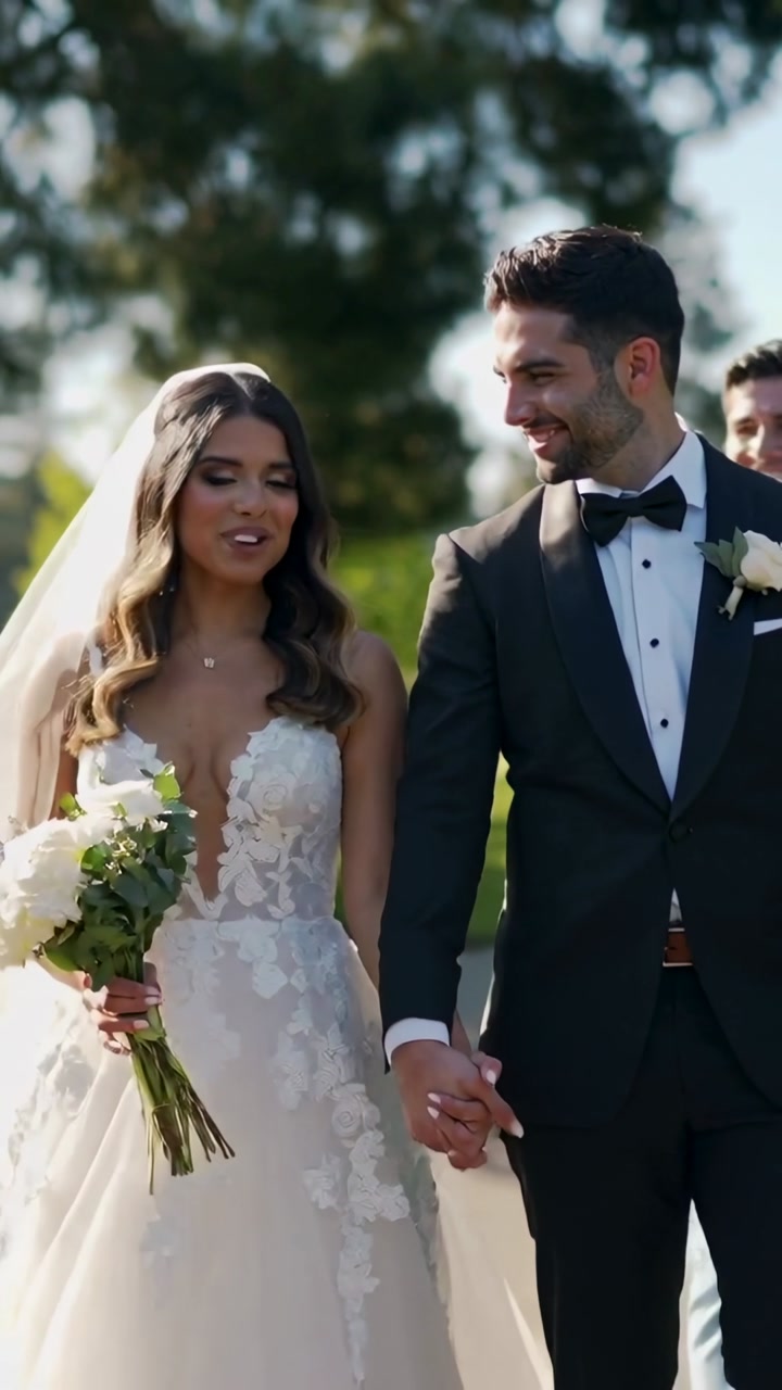 Vertical video: Walking bride in gown, groom in tuxedo holding hands on path after vows, bouquet