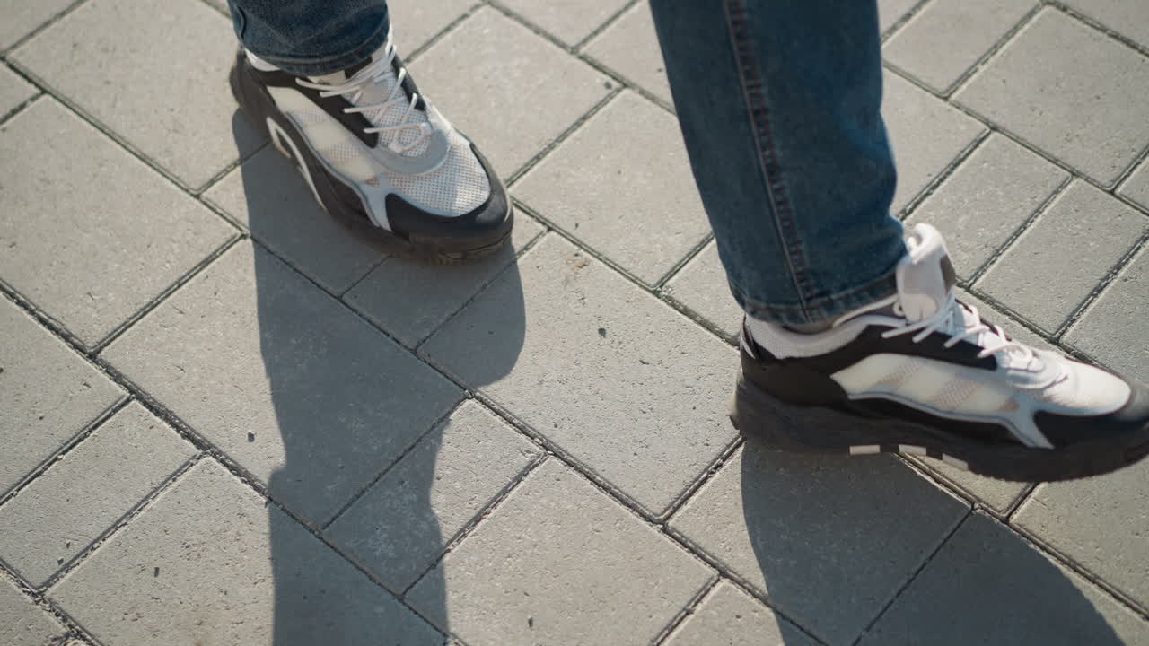 Leg view of person in jeans and black-white canvas sneakers stepping forward on sunlit interlocked pavement tiles with elongated shadow cast on clean surface during clear da