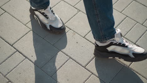 leg view of person in jeans and black-white canvas sneakers stepping forward on sunlit interlocked pavement tiles with elongated shadow cast on clean surface during clear da