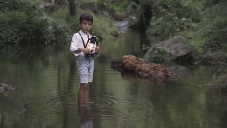 a child standing alone in a river in the middle of the forest holding a lamp or a lantern and wearing vintage clothes