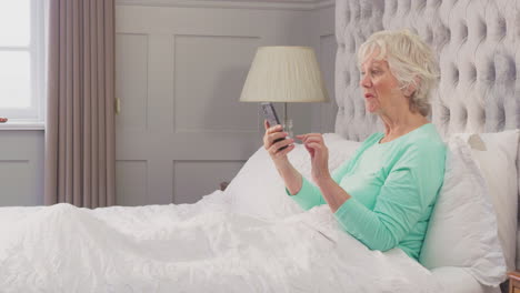 Smiling-Senior-Woman-At-Home-Wearing-Pyjamas-In-Bed-Using-Mobile-Phone