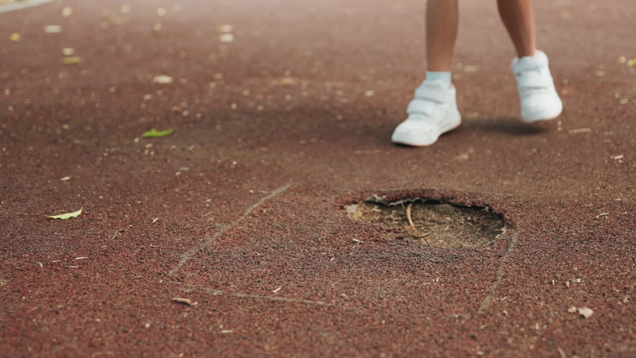 Bambina che si avvicina a un buco nella superficie del parco giochi, scarpe da ginnastica bianche che graffiano la superficie antitrauma in gomma, altalena a fune intrecciata in alto, dita curiose che testano la consistenza, potenziale preoccupazione per la sicurezza, momento spontaneo di gita al parco.
