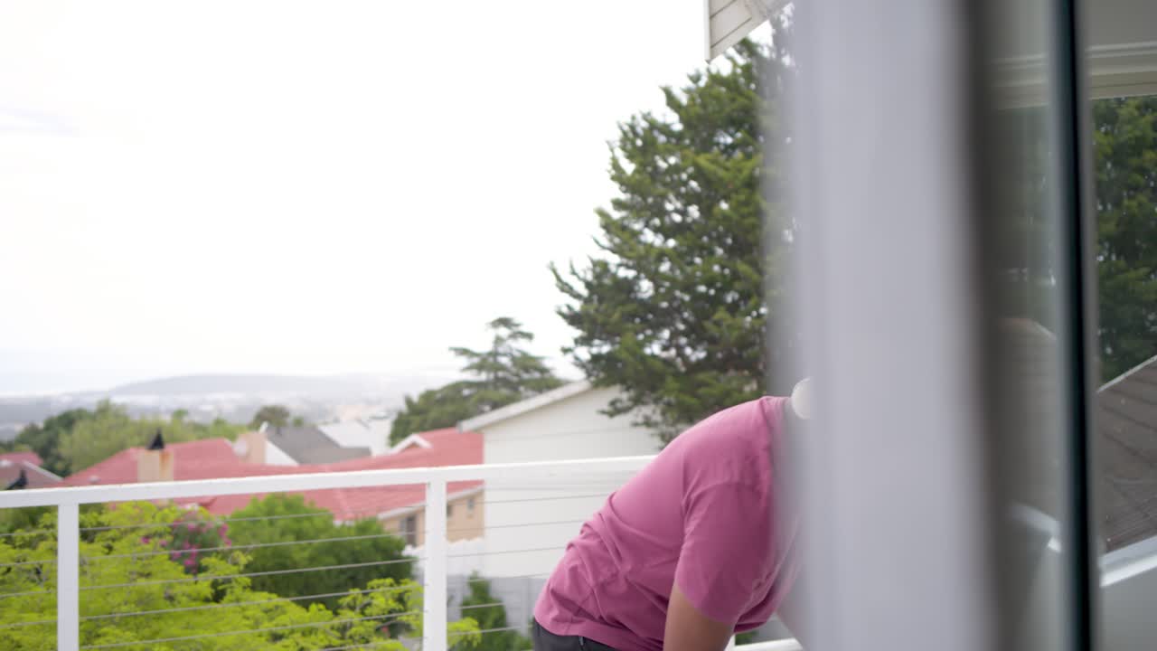 Uomo che spruzza da una bottiglia blu e pulisce le porte scorrevoli sul balcone, piegandosi per ispezionare, nell'ambito delle pulizie di casa.